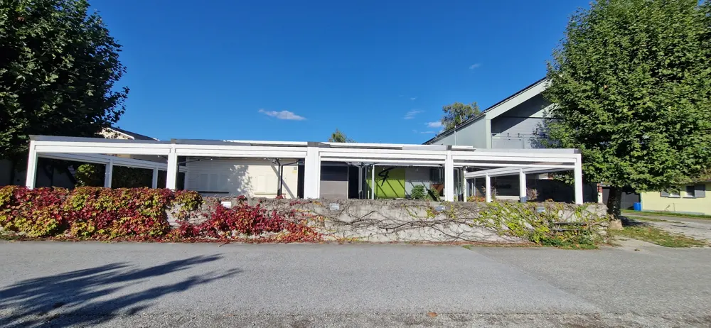 Exterior view of a white pergola with climbing vines on a stone wall under a blue sky.