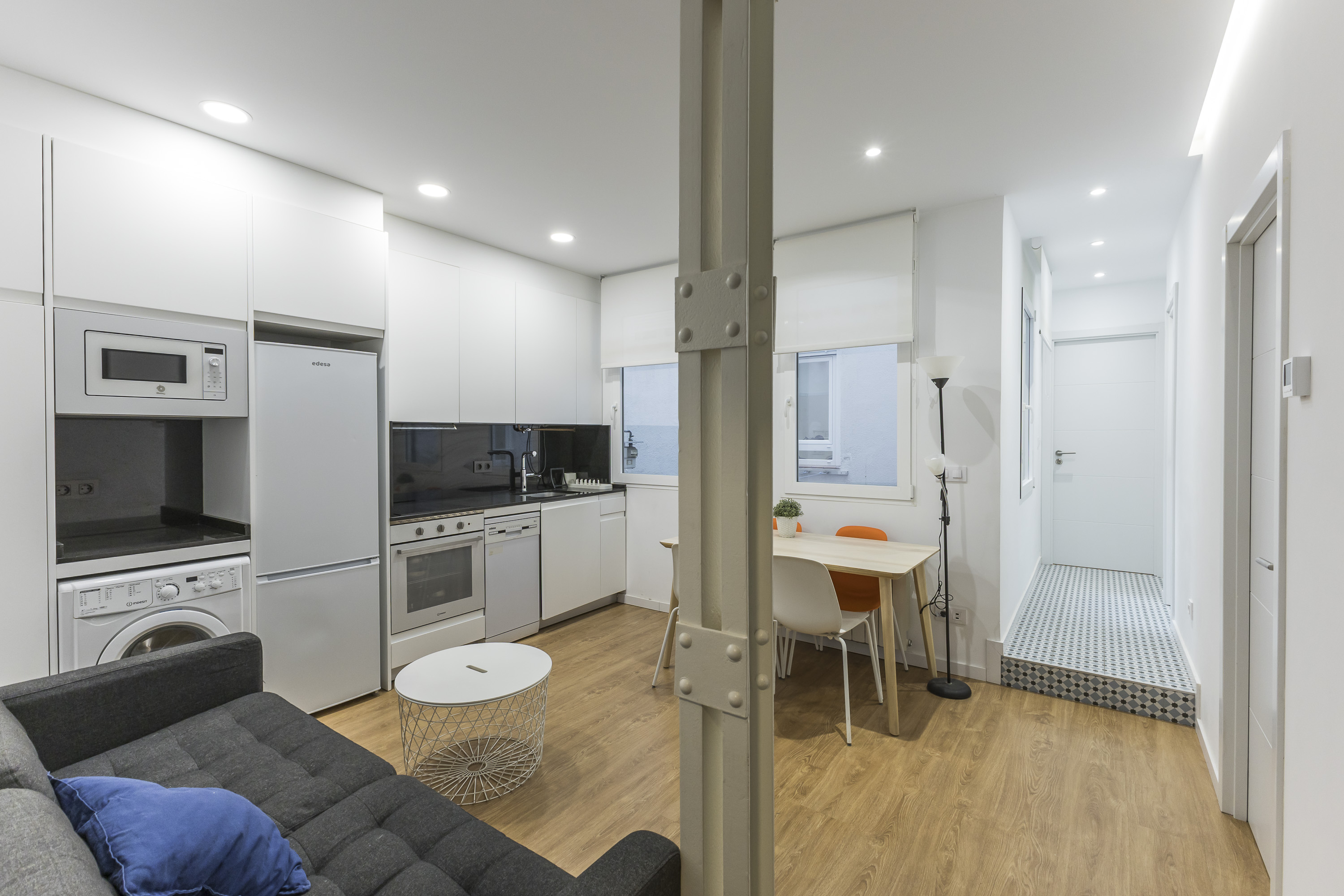 Modern open-plan apartment with a grey sofa, white kitchen, dining area, and wood floors, divided by a structural pillar. [AI-generated text]