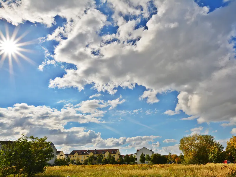 Sunny sky with clouds over a field of tall grass and buildings. The sky is blue with white clouds and a bright sun.