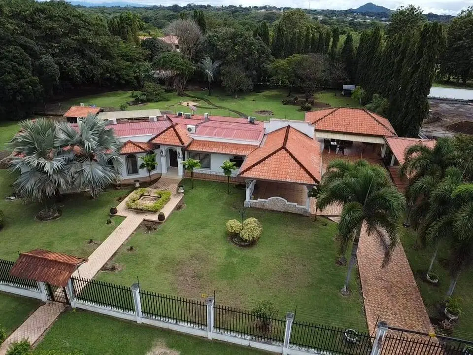 Aerial view of a white house with a red tile roof, surrounded by green lawn, trees, and a black fence.