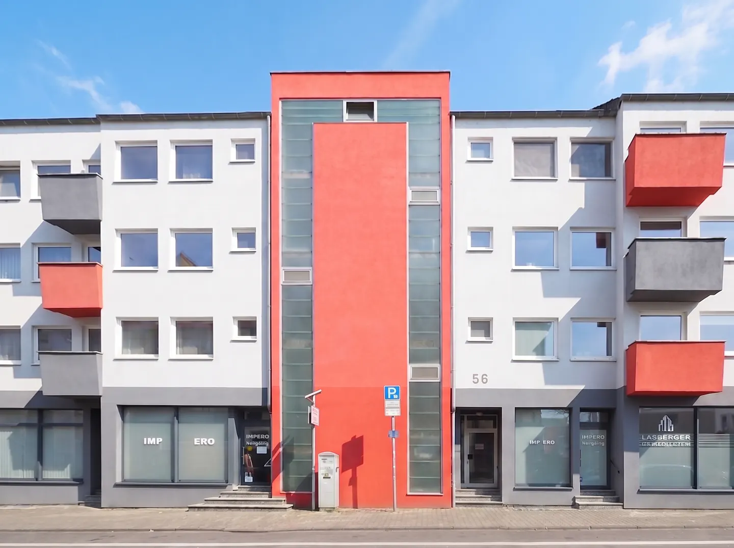 Facade of a three-story white building with red accents and glass panels under a blue sky.