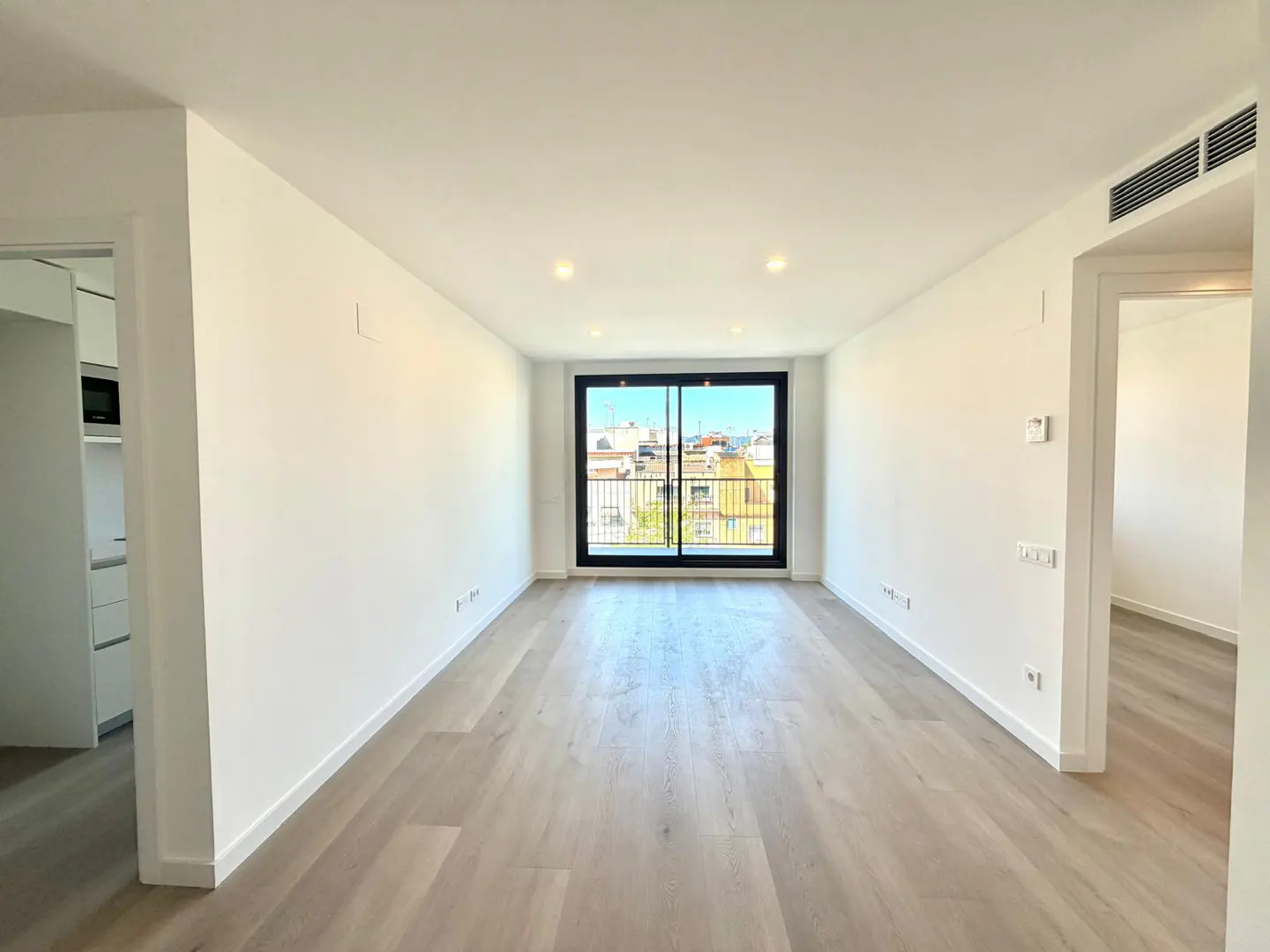 Bright, empty room with light wood floors, white walls, and a black-framed sliding glass door to a balcony.