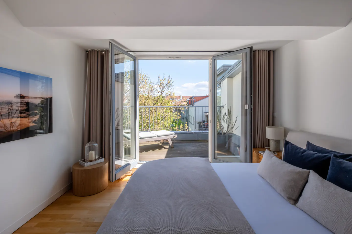 Bedroom with white walls, wood floor, and open doors to a balcony with a lounge chair and plants.