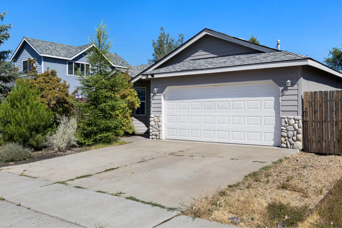 A gray single-story house with a white garage door and stone accents on a sunny day. A blue house is visible in the background.