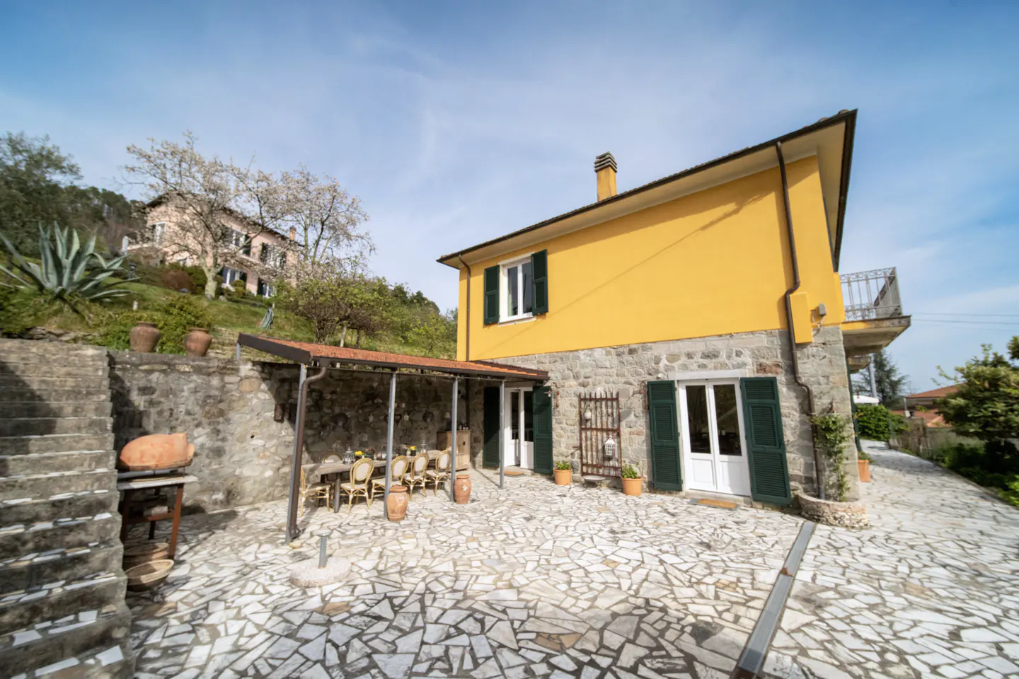 Exterior view of a yellow house with stone accents, green shutters, and a stone patio with outdoor seating.