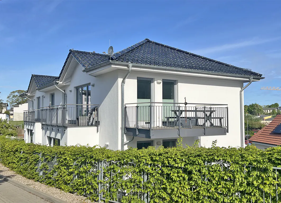 A two-story white house with a dark roof and balconies, surrounded by a green hedge.