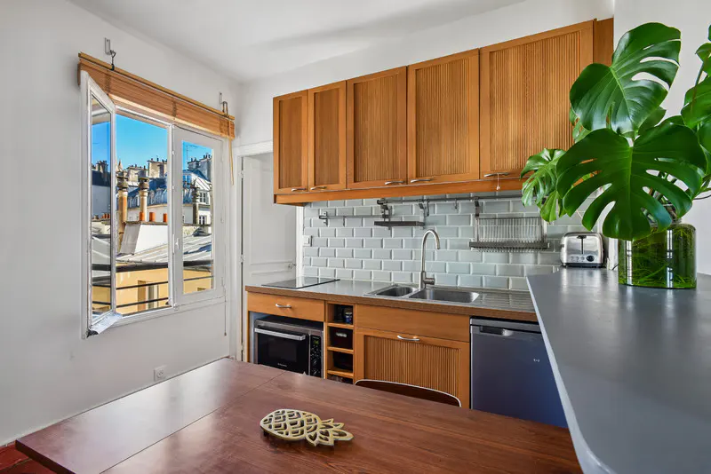 Bright kitchen with wood cabinets, subway tile backsplash, and a view of Paris rooftops through an open window. A dark wood table sits in the foreground.