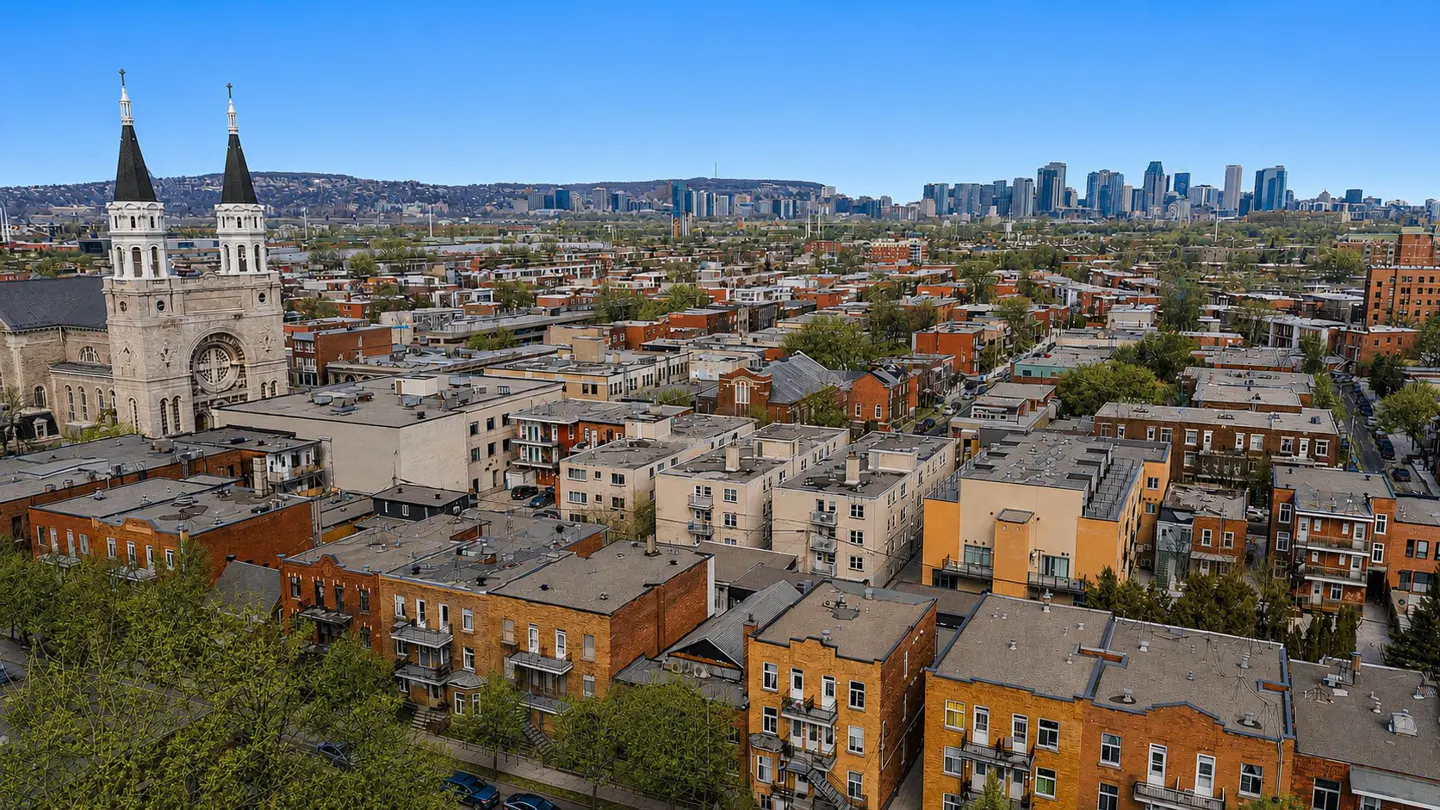 Aerial view of a dense urban neighborhood with brick buildings, a church with tall spires, and a city skyline in the distance under a clear blue sky.