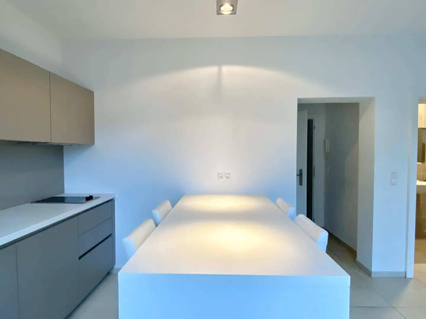 Modern kitchen with gray cabinets, white countertops, and a large white table with chairs. A doorway leads to another room.