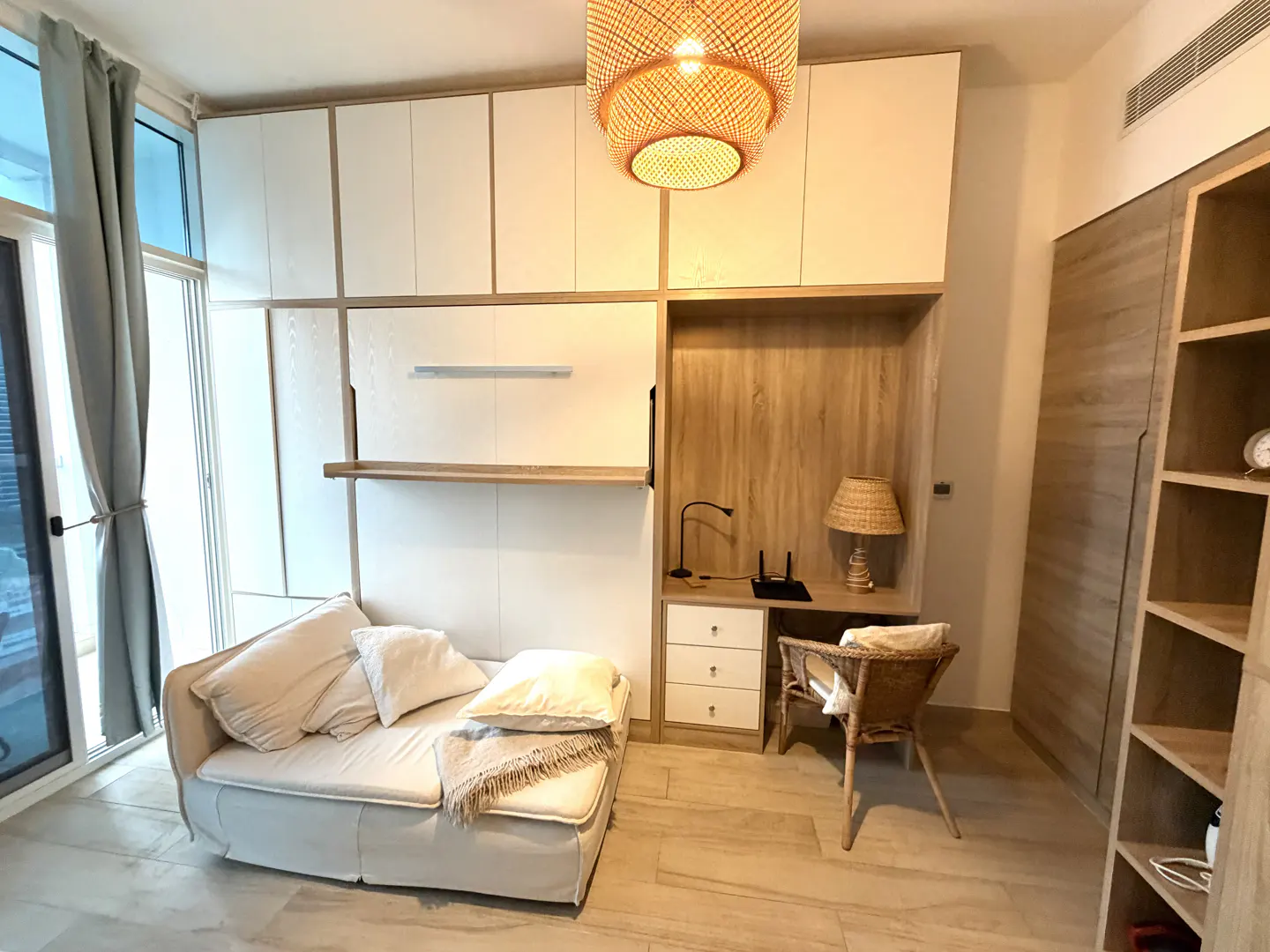 A bright room with a white sofa, pillows, and a woven blanket. A desk with a lamp and chair are built into the wall unit. A woven light fixture hangs from the ceiling.