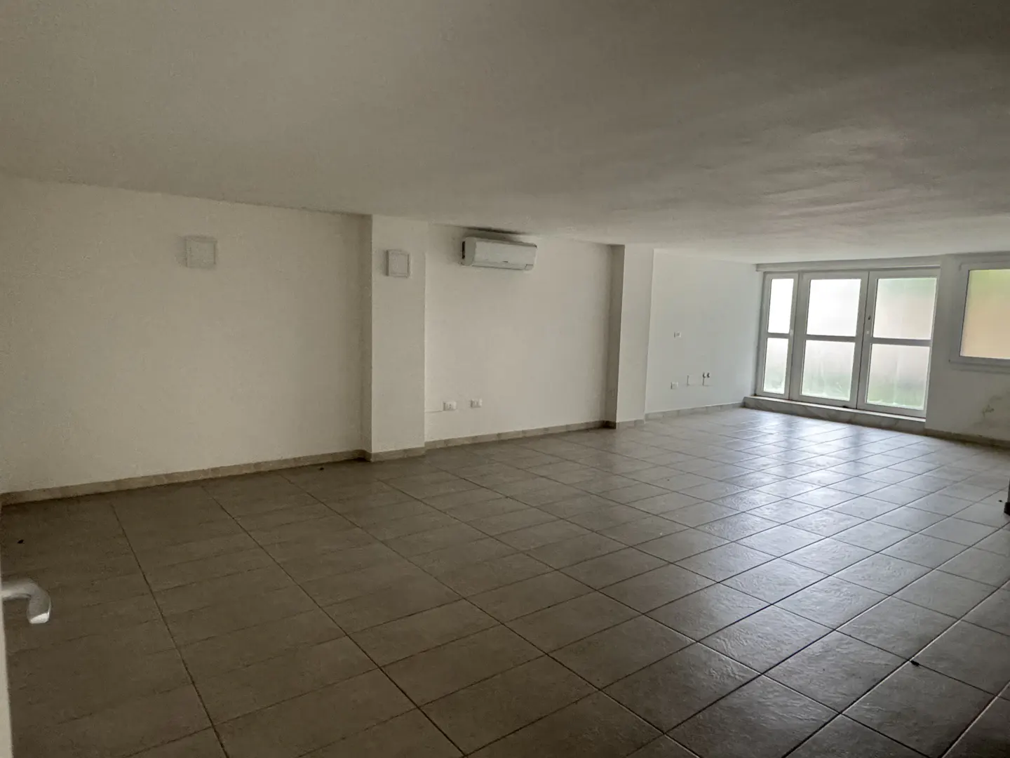Empty, spacious room with white walls, beige tiled floors, an air conditioning unit, and large windows.