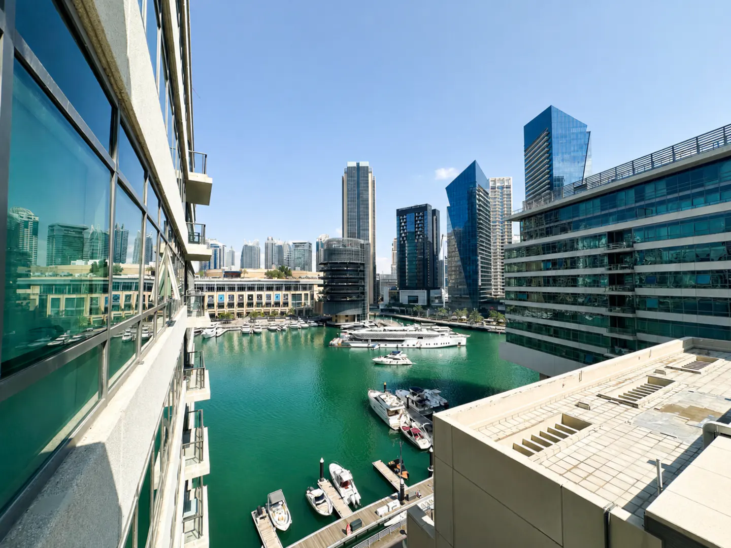 View from a balcony overlooking a turquoise marina with docked yachts and modern glass skyscrapers under a clear blue sky.