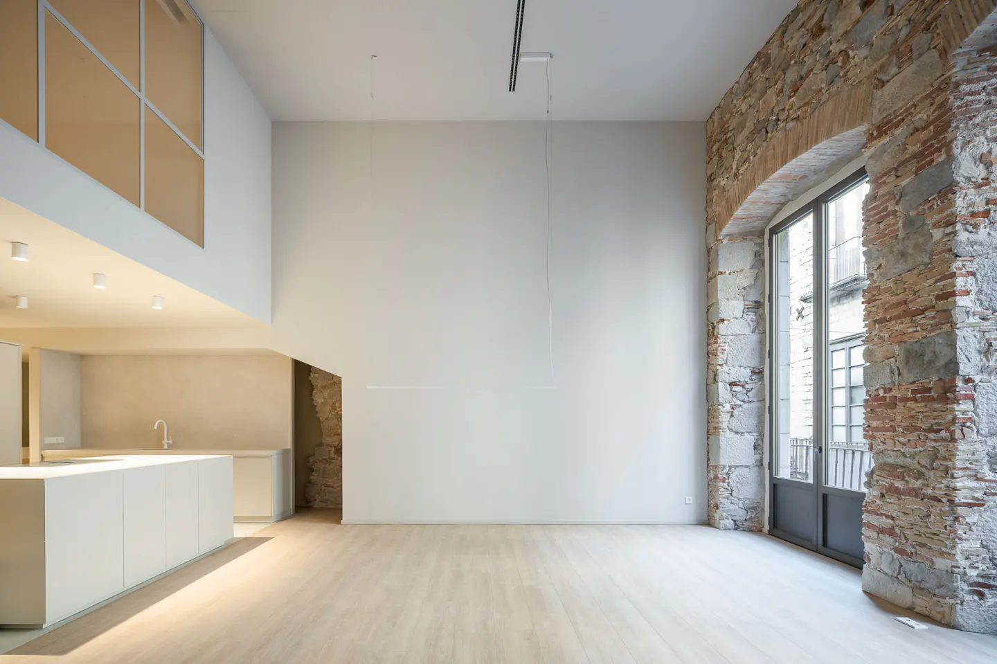 Bright, modern apartment interior with white walls, light wood floors, and exposed brick around a large window. Kitchen area visible.