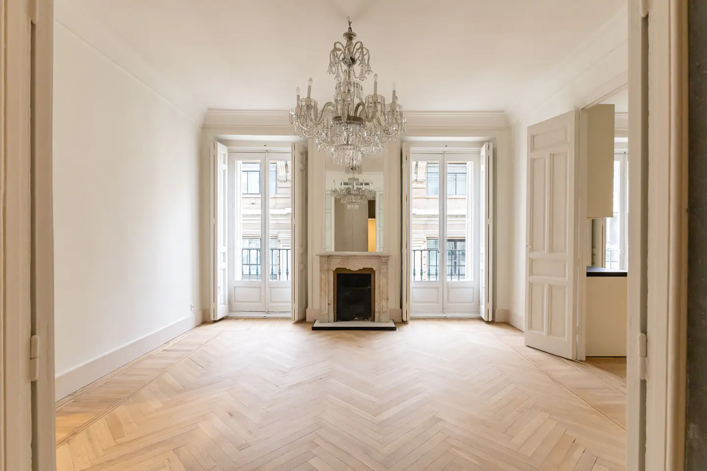 Bright, empty room with herringbone wood floors, white walls, and a crystal chandelier. Two sets of French doors flank a marble fireplace.