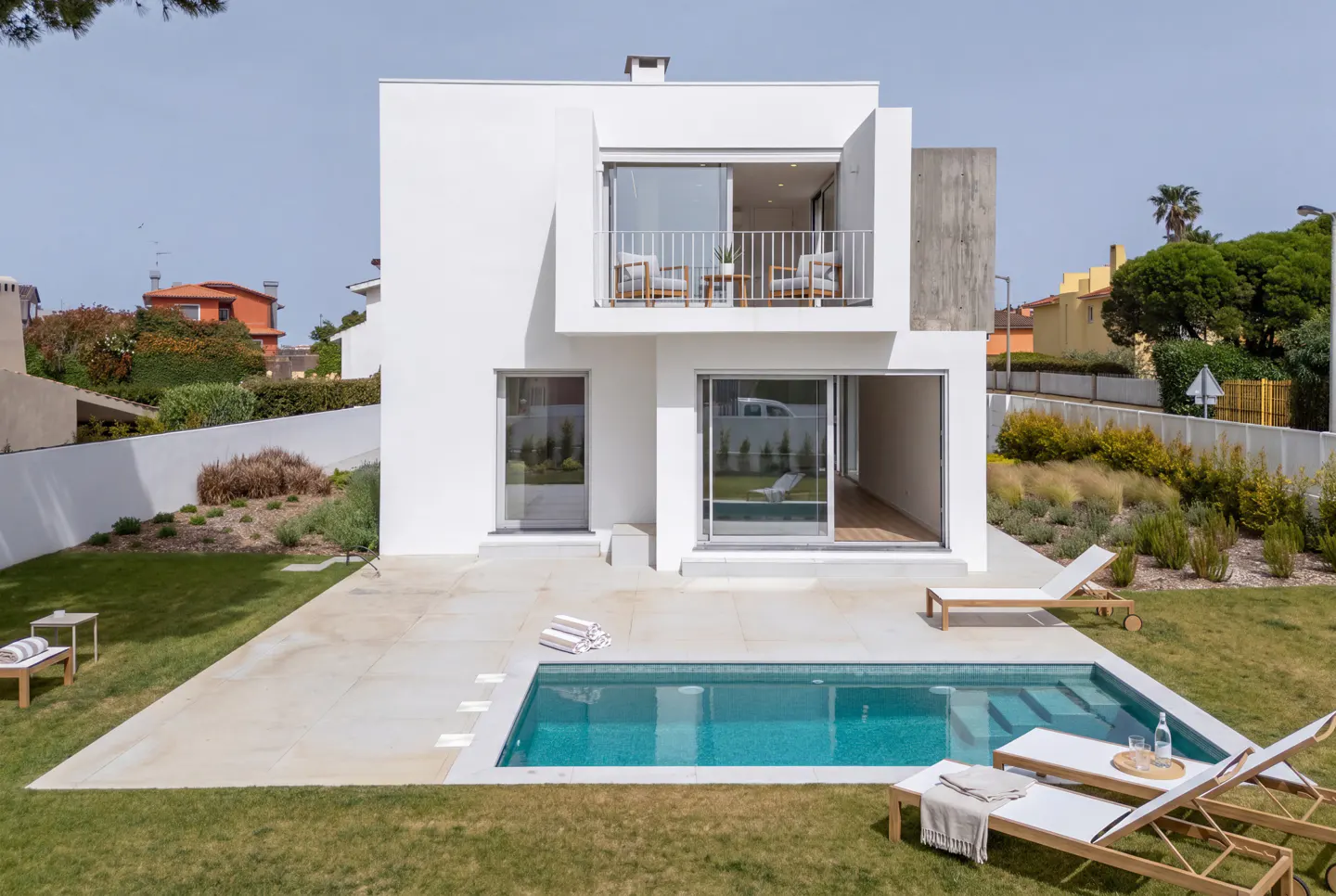 Modern two-story white house with a balcony overlooking a turquoise pool and lawn. Lounge chairs are poolside.