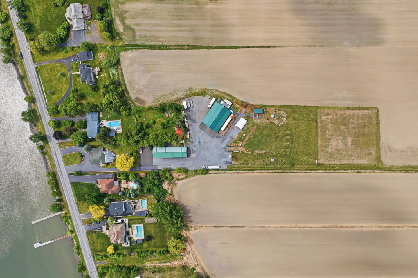 Aerial view of rural properties along a river, featuring houses with pools, a green-roofed barn, and large open fields.