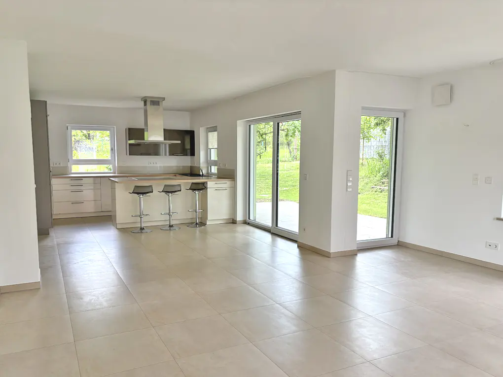 Bright, open-plan kitchen and living area with beige tile flooring. Kitchen island with three stools, sliding glass doors to a green yard.