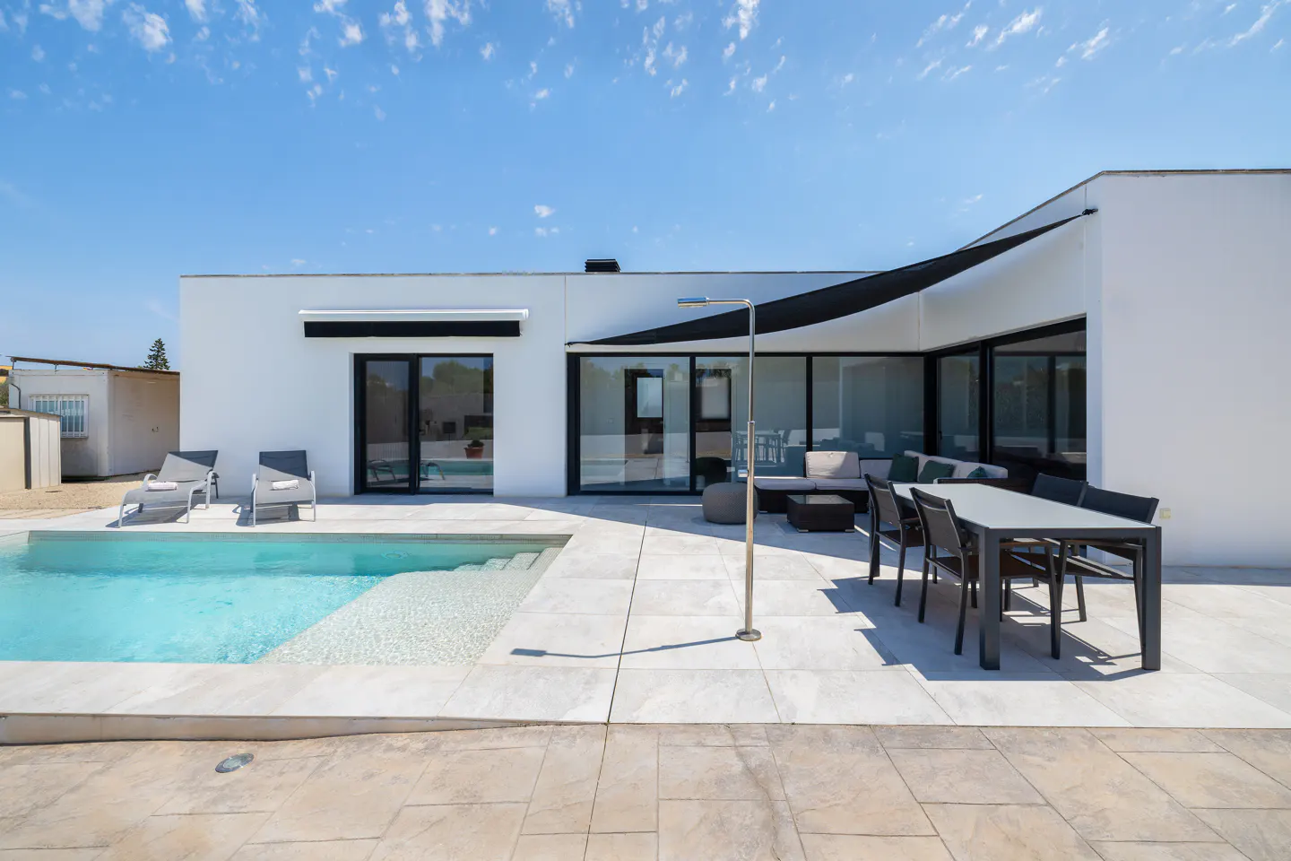 Modern white villa with a swimming pool, patio dining set, lounge chairs, and a black sun shade under a clear blue sky.