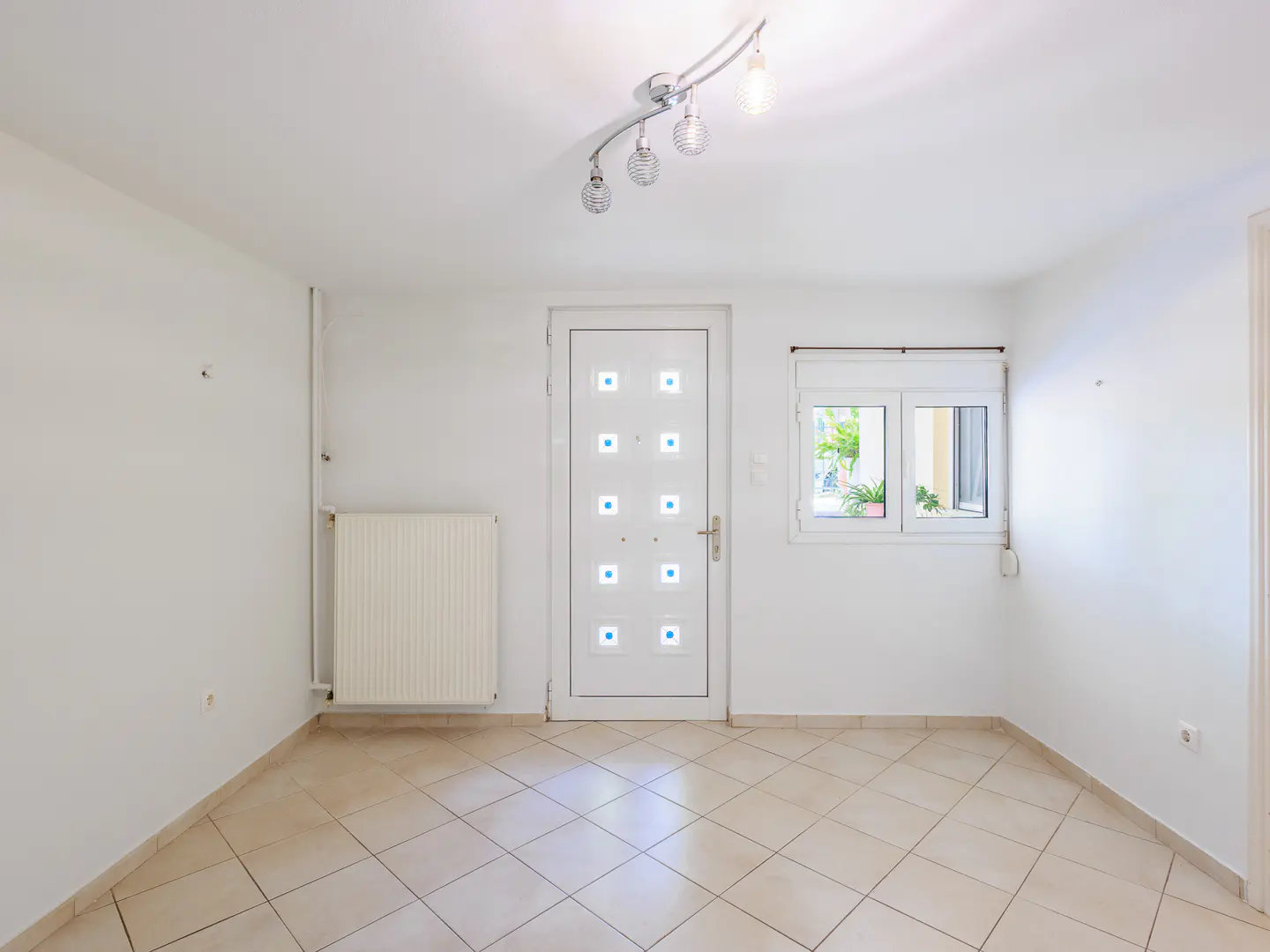 Bright, empty room with white walls, tiled floor, radiator, and a white door with blue squares. A window shows greenery outside.