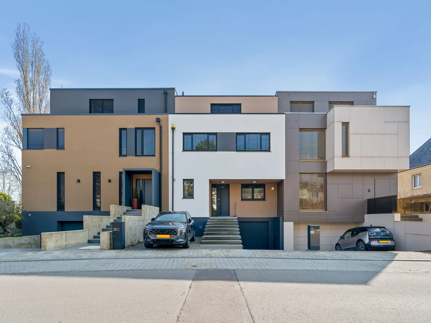 Modern townhouses in beige, white, and gray, with cars parked in front, under a clear blue sky.