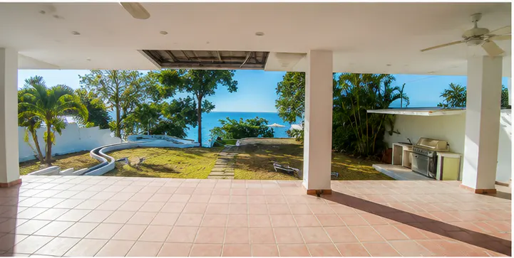Covered patio with terracotta tile floor, white pillars, and a view of a green lawn, trees, and the ocean. A grill is visible on the right.
