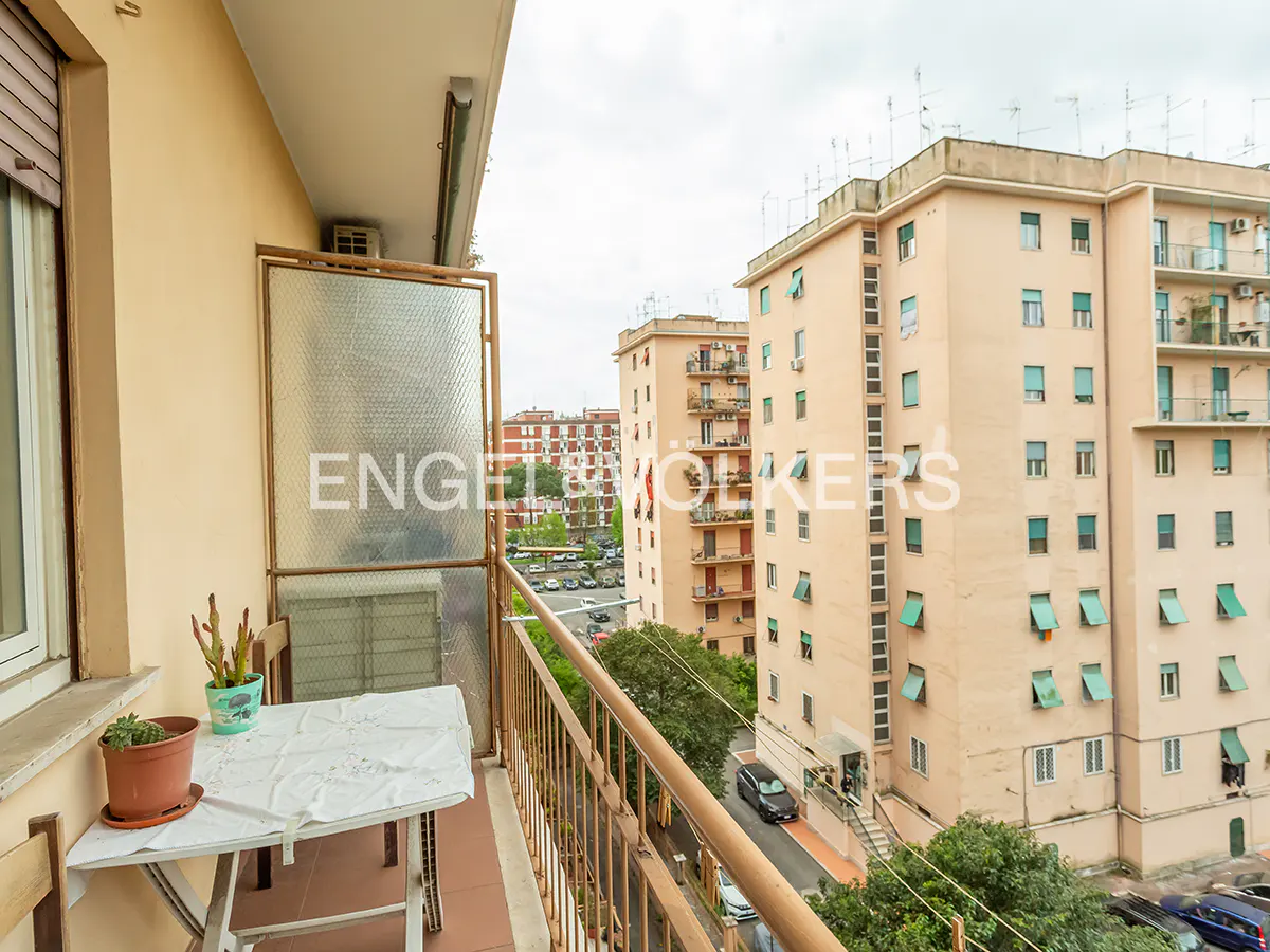 Balcony view with a small table, potted plants, and a metal railing overlooking city buildings on a cloudy day.