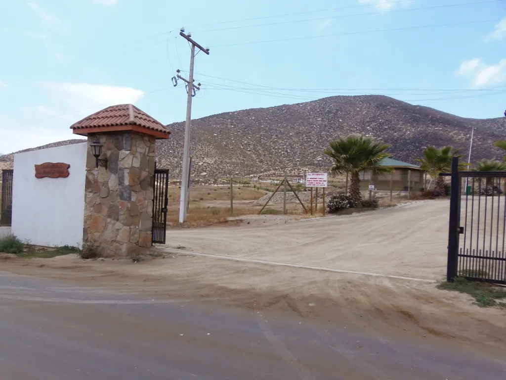 Gated entrance to a property with a stone pillar, white wall, and a sign. A dirt road leads to a building with palm trees.