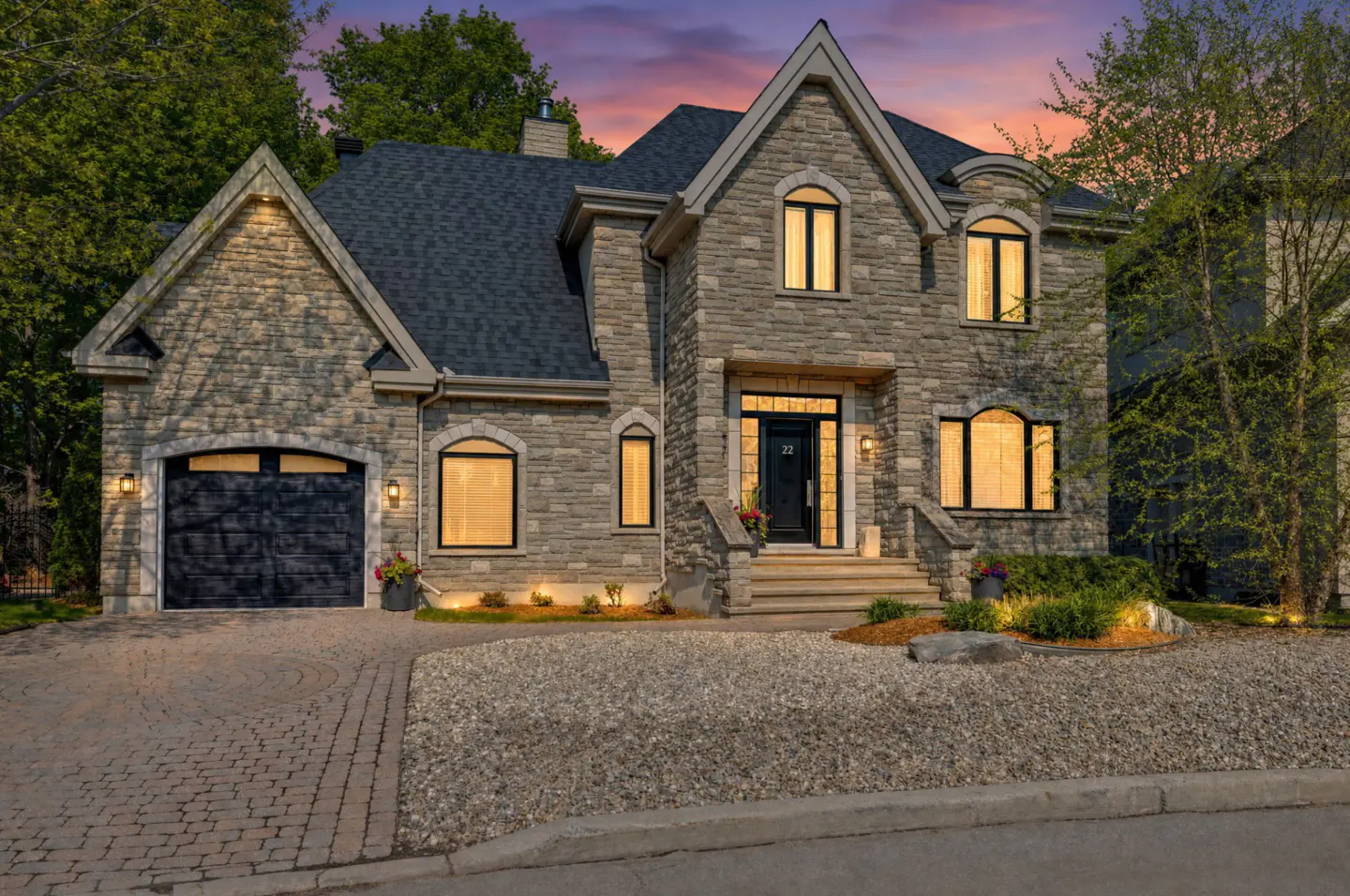 Two-story stone house with a black garage door, black front door, and a dark gray roof. The yard is gravel with some greenery.