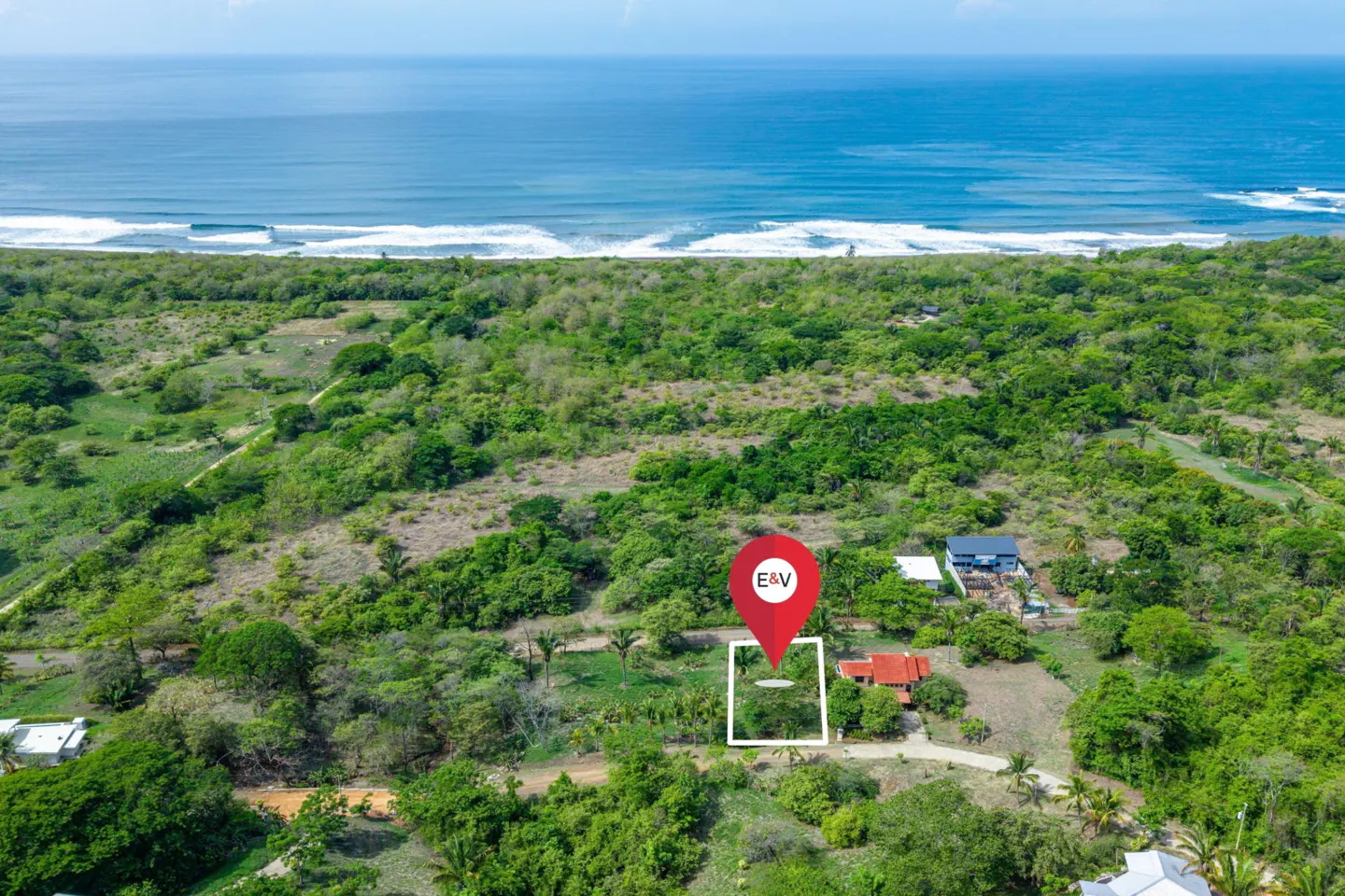 Aerial view of a property near the ocean, marked with a red pin and white square. Lush green trees surround the lot.