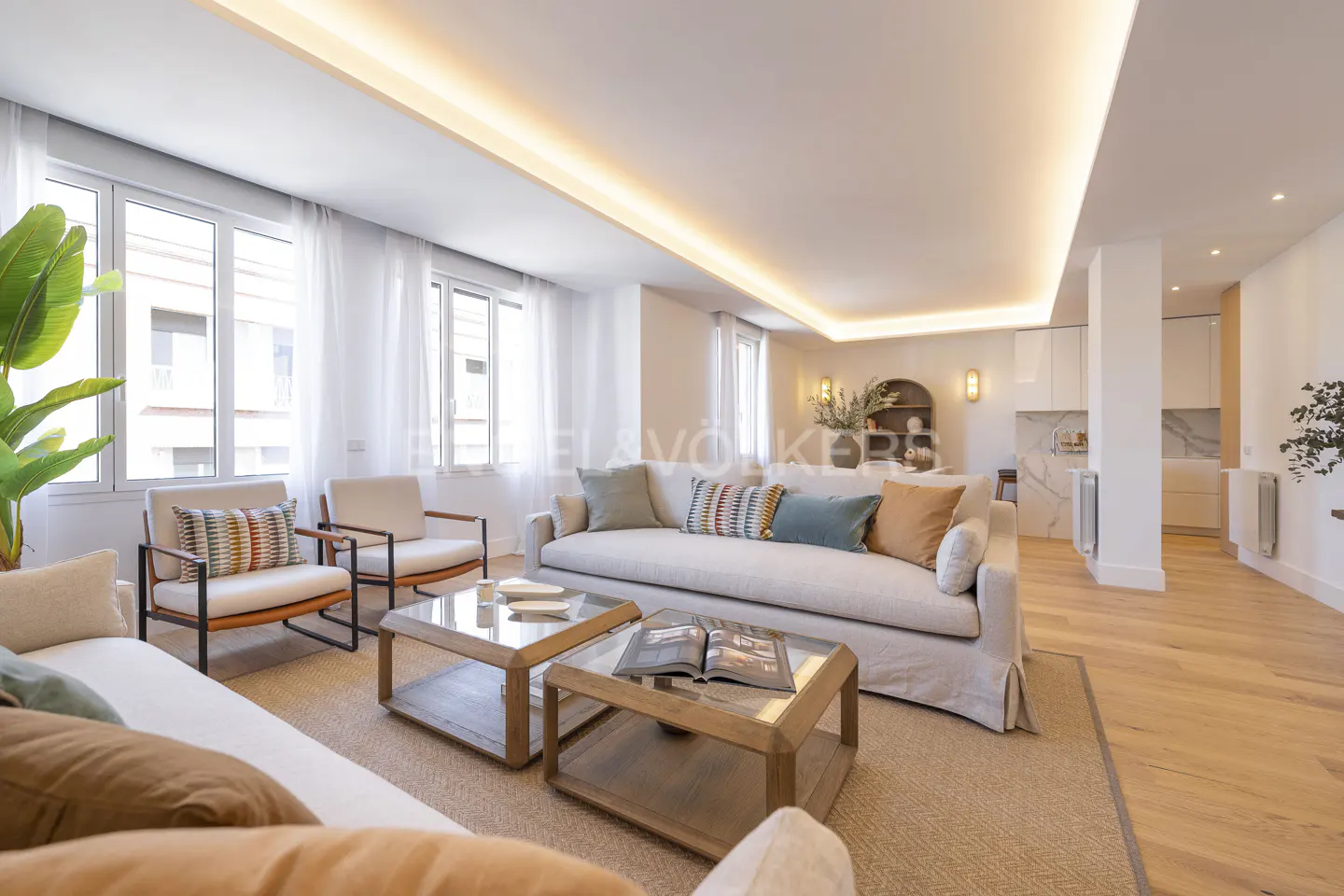 Bright living room with white sofas, armchairs, and glass coffee tables on a jute rug. Large windows with sheer curtains and recessed lighting. Open to a white kitchen.