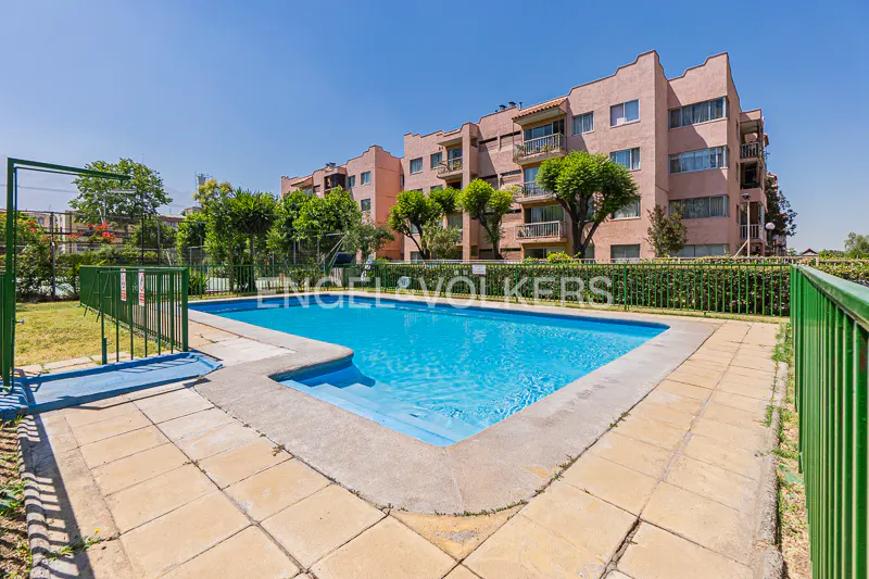 Outdoor pool with turquoise water, surrounded by tan tiles and a green fence. A pink apartment building is in the background.