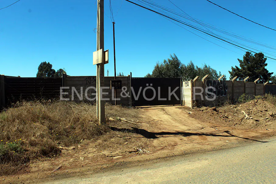 Gated property entrance with Engel & Völkers logo, concrete walls, dirt road, and dry vegetation under a clear blue sky.