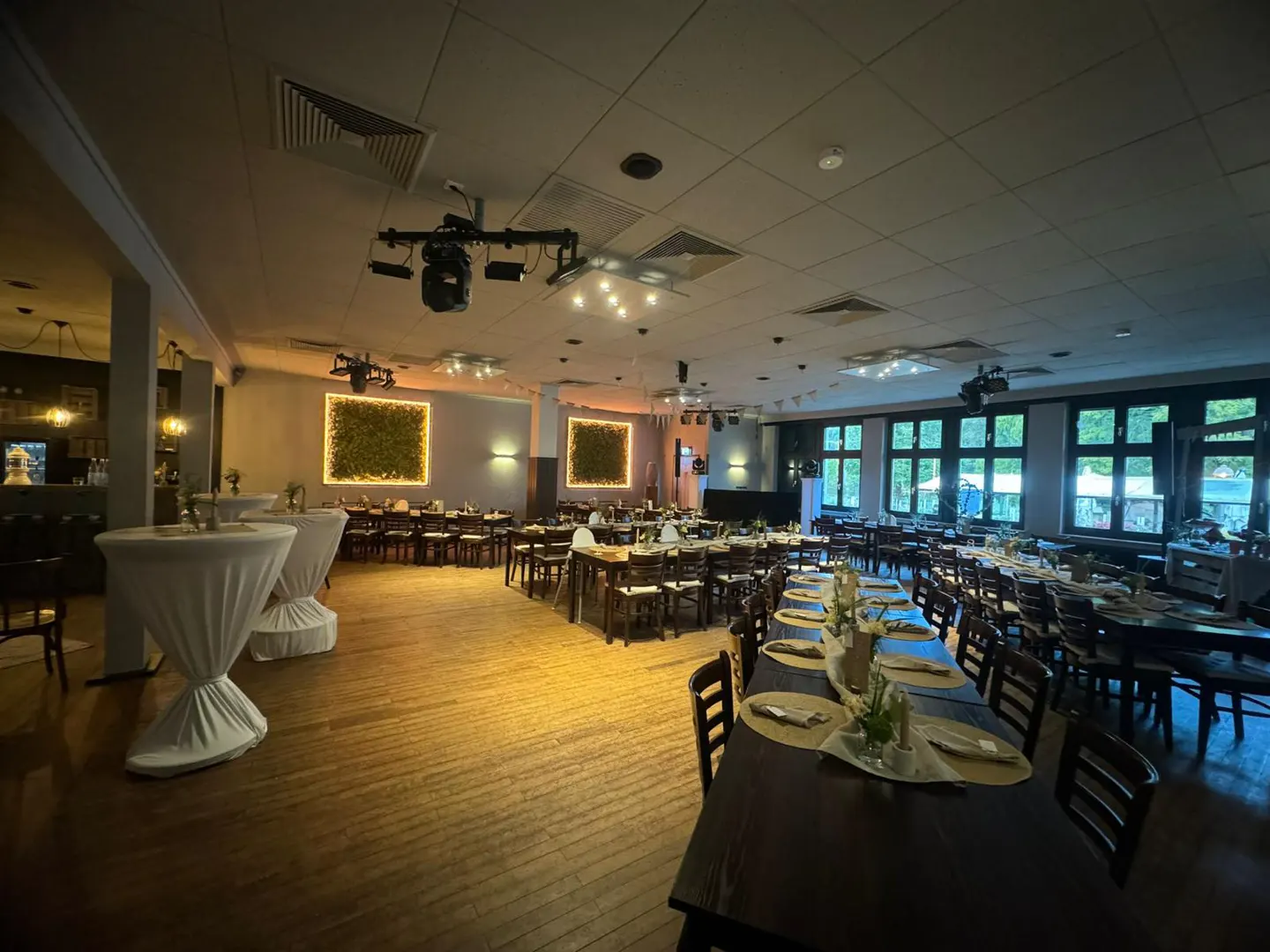 Spacious event hall with wooden floors, long dining tables, white-covered cocktail tables, and decorative wall lighting.