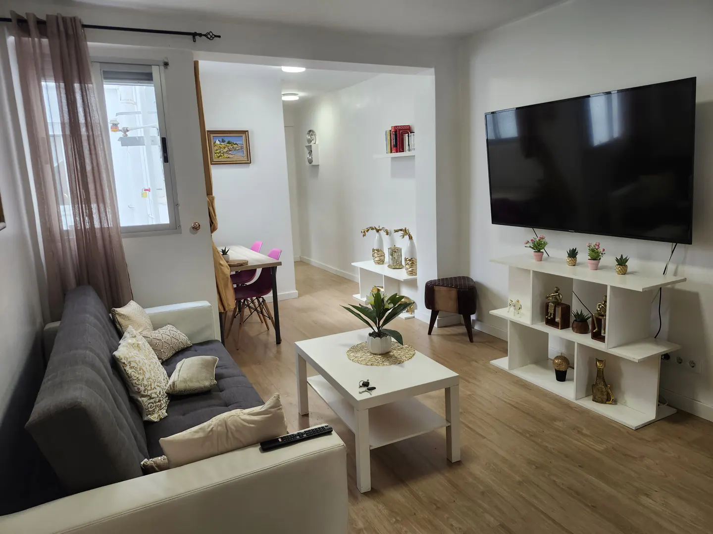 Modern living room with a grey sofa, white coffee table, wall-mounted TV, and a dining area visible in the background.