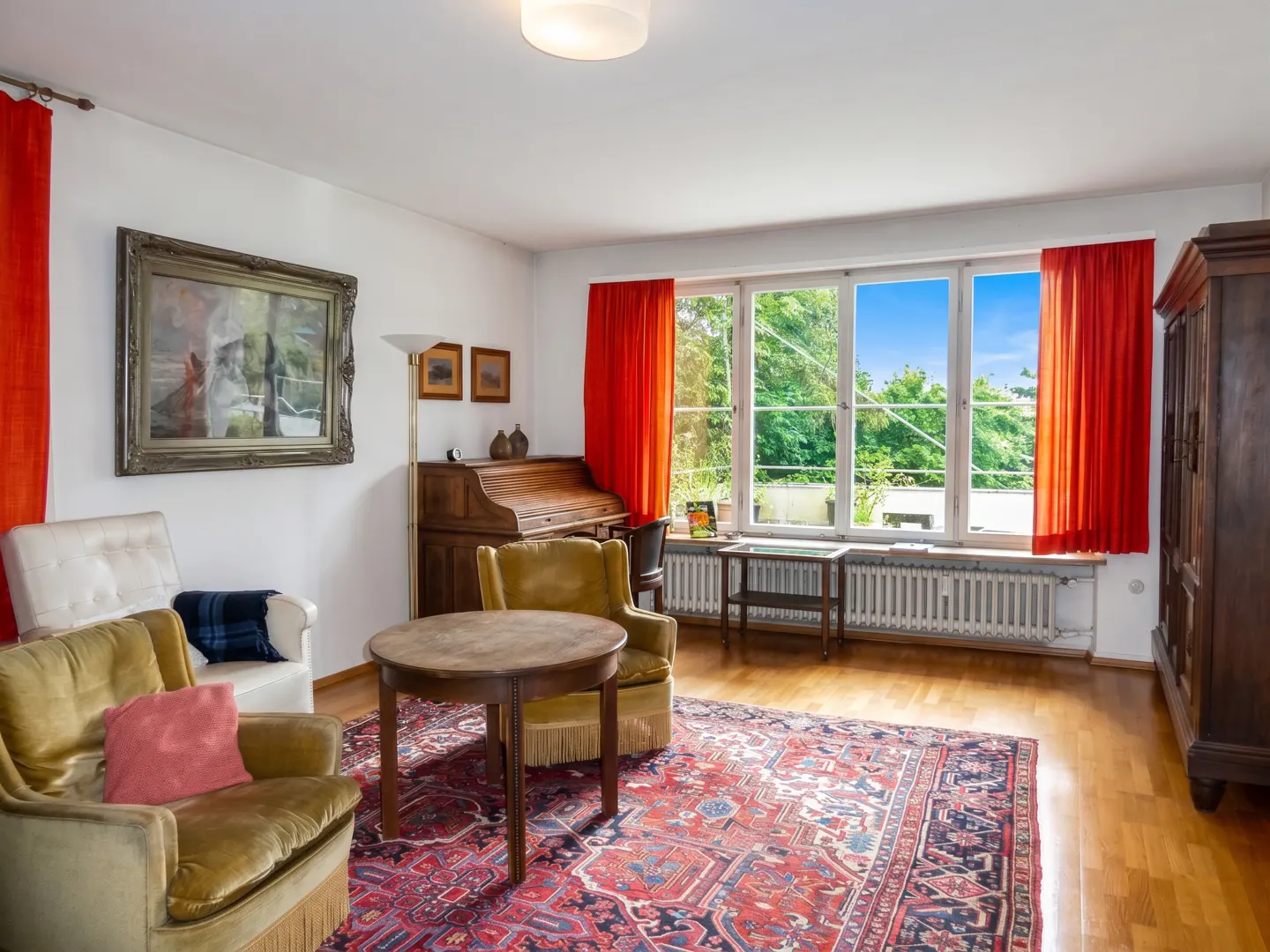 Living room with vintage armchairs, a round wooden table, a patterned rug, and large windows with bright orange curtains.
