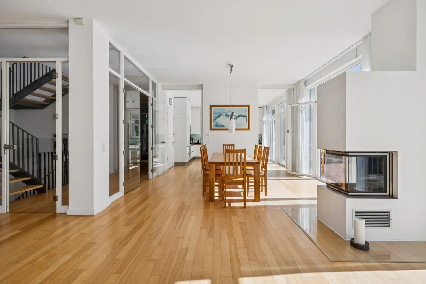 Bright, modern dining room with wood floors, white walls, and a fireplace. A wooden table and chairs sit under a framed painting. Glass doors lead to a staircase.