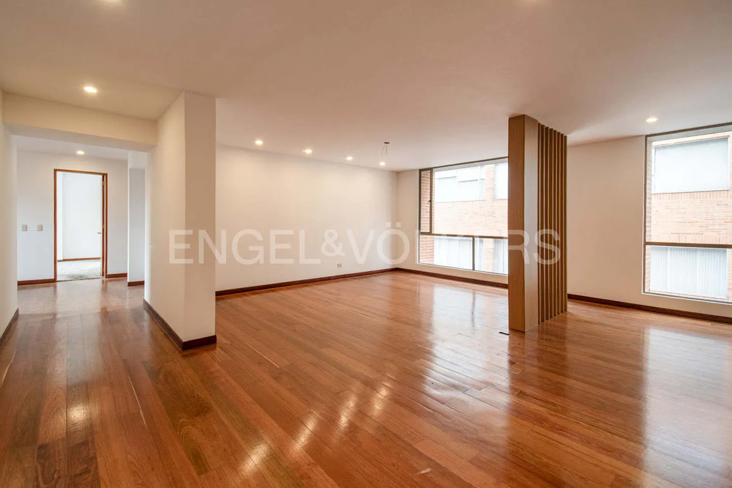 Bright, empty room with hardwood floors, white walls, and two large windows. Engel & Völkers logo visible.