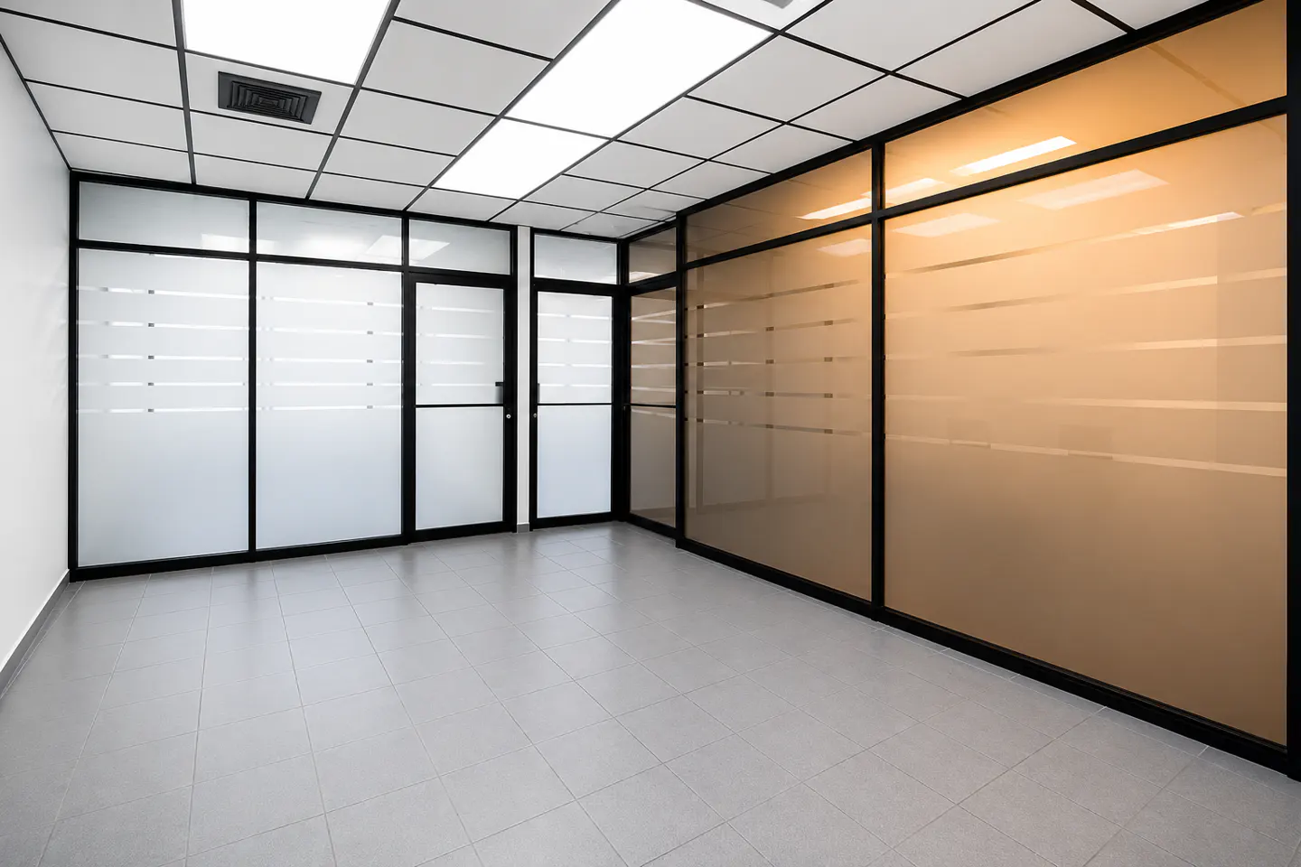 Empty office space with grey tiled floors, drop ceiling, and glass partitions featuring frosted horizontal stripes.