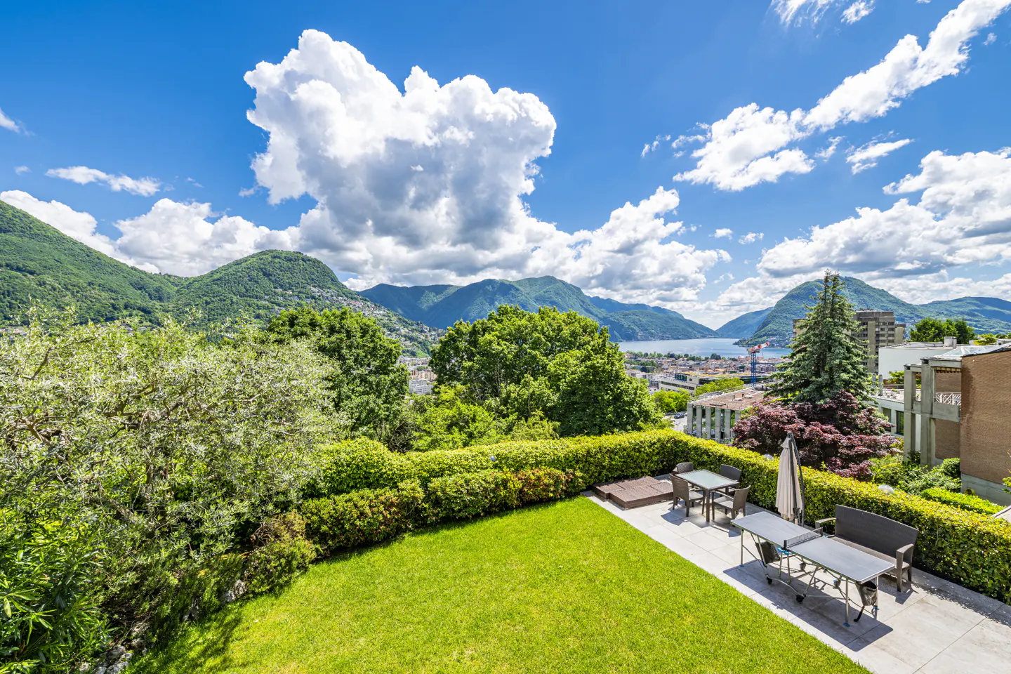 Scenic view from a green lawn with patio furniture, overlooking mountains and a lake under a blue sky with white clouds.