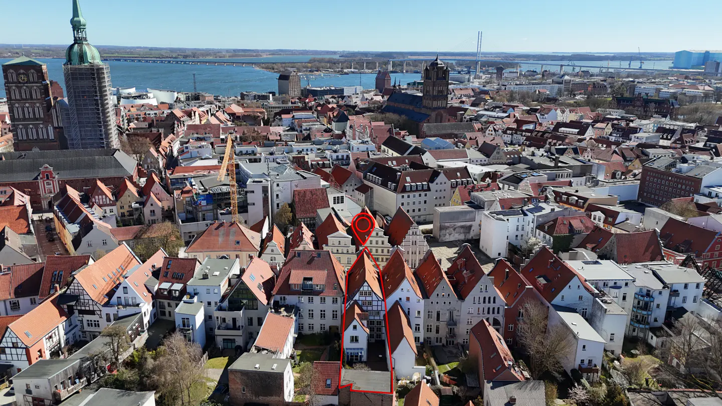 Aerial view of a European city with red-tiled roofs, tall church spires, and a river in the background. A red outline highlights a property.
