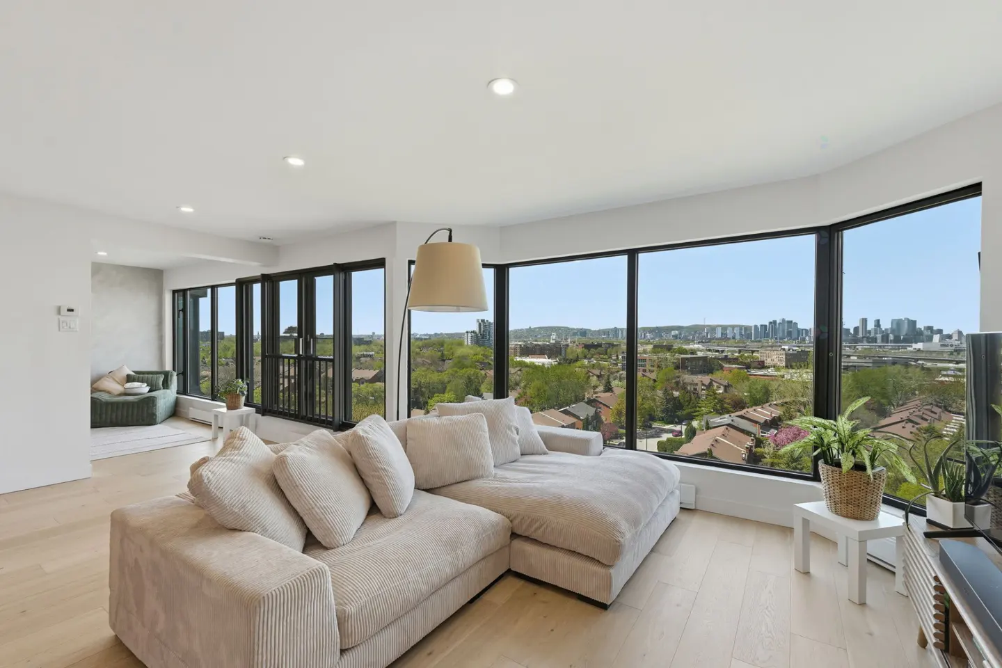 Bright living room with a large beige sectional sofa, floor lamp, and floor-to-ceiling windows with a city view.