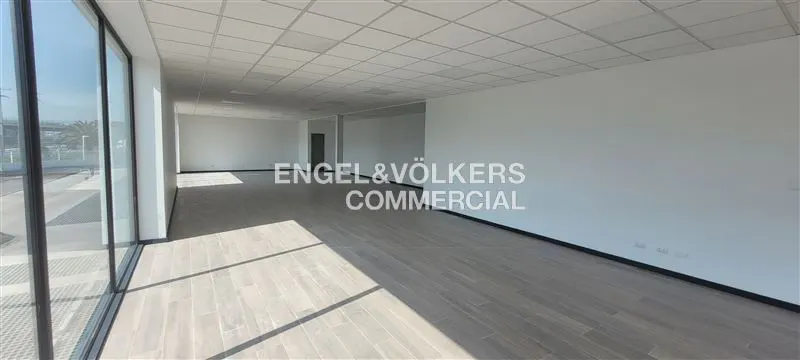 Bright, empty commercial space with wood floors, white walls, and large windows. "Engel & Völkers Commercial" logo on the wall.