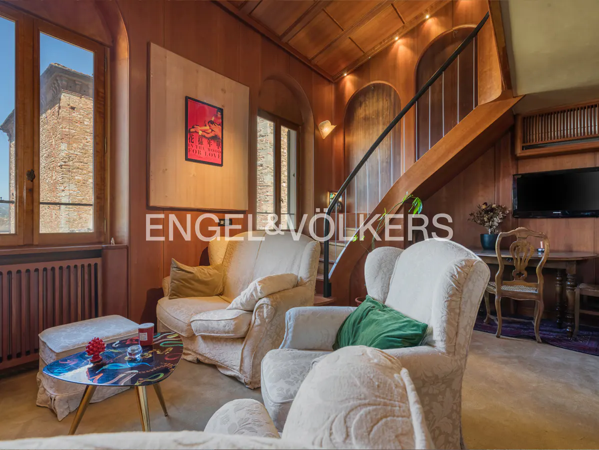 Living room with wood paneled walls, cream armchairs, and a staircase. A window shows a stone building outside.