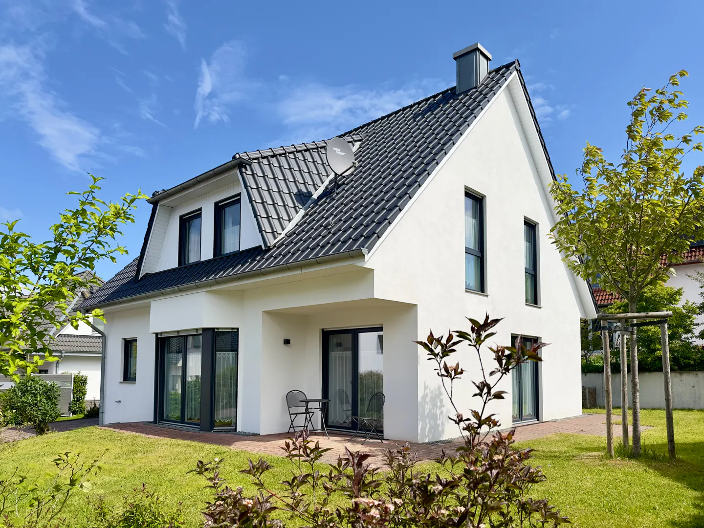Modern white house with a dark gray tiled roof, green lawn, and blue sky. A small patio has a table and chairs.