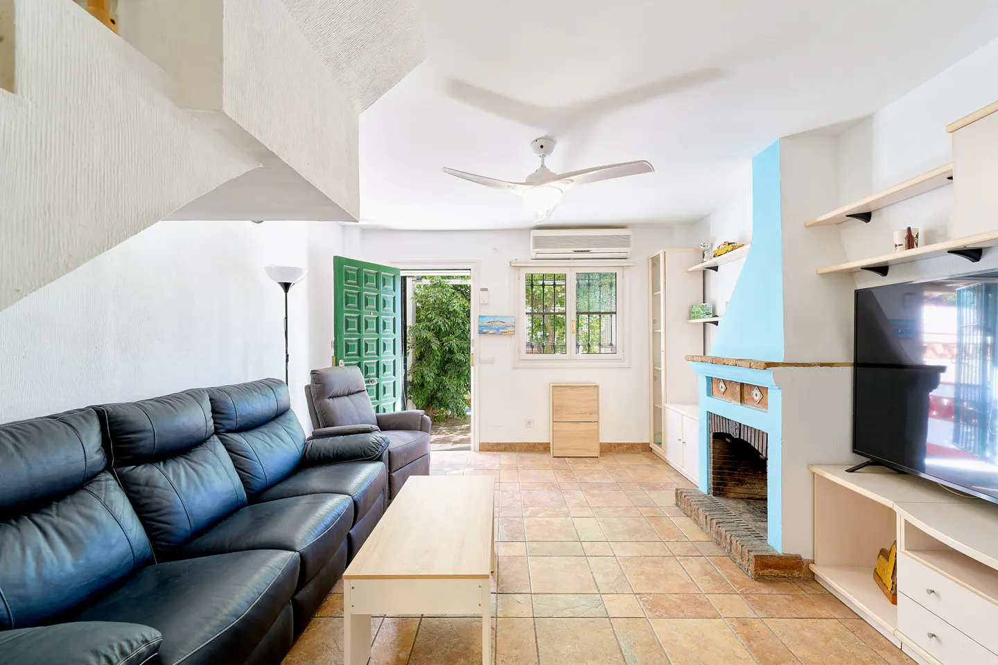 Living room with a black leather sofa, a light wood coffee table, and a green door leading to a garden. The room has tile floors and a blue fireplace.