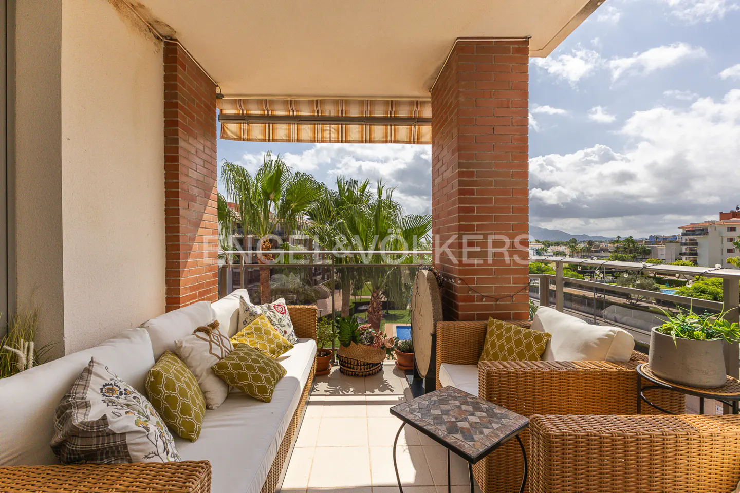 Balcony with wicker furniture, white cushions, and a mosaic table. Palm trees and city views are visible in the background.