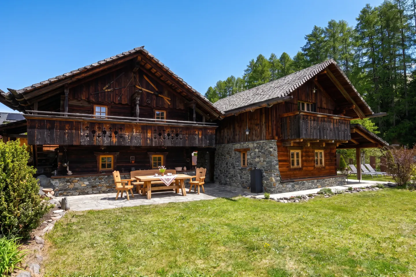 Exterior view of a two-story wooden house with a stone foundation and a wooden table with chairs on the patio.