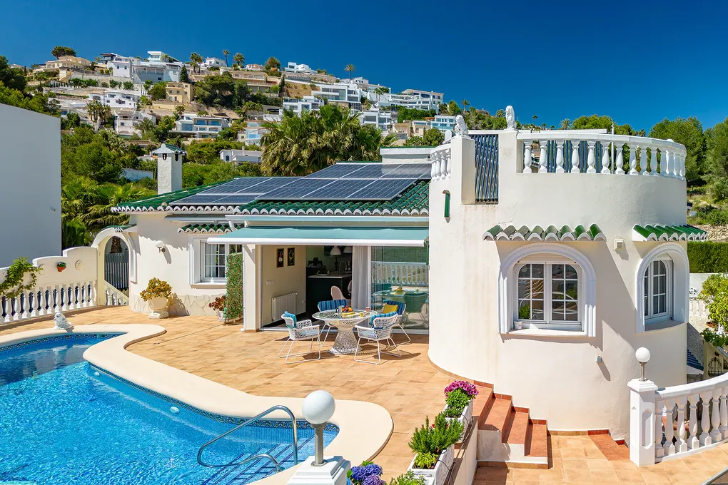 Exterior view of a white villa with a pool, terrace, and solar panels on the roof, set against a hillside of white houses under a blue sky.