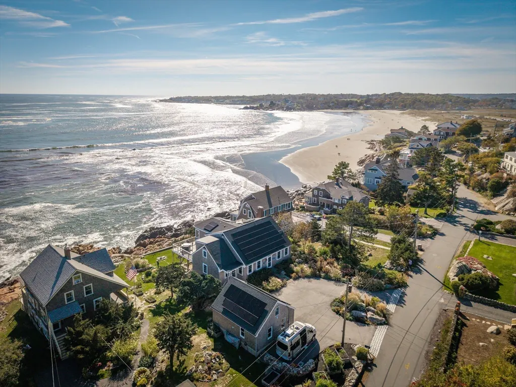 Aerial view of coastal homes with solar panels, a sandy beach, and the ocean under a blue sky.