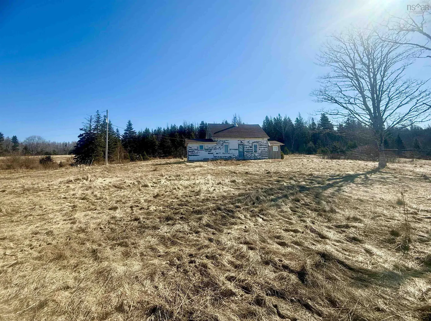 Exterior view of a weathered, two-story white house in a field of dry grass under a clear blue sky. Trees line the background.